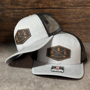May include: Two heather gray trucker hats with black mesh backs and adjustable snapback closures. Each hat features a brown leather-like patch with the letters M and K, an ampersand, and the date 04.25.2026 engraved with crossed arrows.