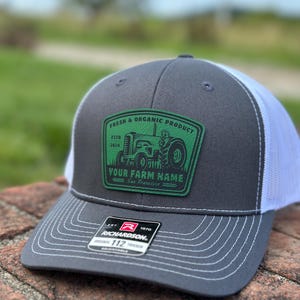 May include: A gray and white trucker hat featuring a green patch with a tractor illustration and the text FRESH & ORGANIC PRODUCT ESTD 2024 YOUR FARM NAME San Francisco. A black Richardson 112 adjustable sticker is on the brim with white stitching.