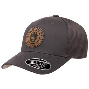 May include: A dark gray baseball cap with a mesh back and a circular brown leather patch on the front. The patch features a graphic of a monkey wearing a hat and the text THE LEATHER PATCH.COM HAT CO. A black sticker on the brim reads 110 FLEXFIT TECH.