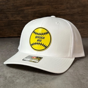 May include: White trucker hat with a yellow softball patch. The patch features the name "BRYLEIGH" and the number "#10" in black text. The hat has a white mesh back and a curved brim.