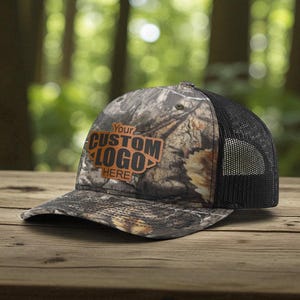 May include: A camouflage print trucker hat featuring a black mesh back and a brown leather-like patch on the front panel. The patch is embossed with the text YOUR CUSTOM LOGO HERE in a bold, block font, centered on the front of the cap.