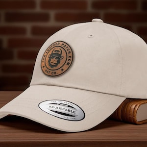 May include: A beige baseball cap with a curved brim. The cap features a circular leather patch with a monkey graphic and the text "THE LEATHER PATCH HAT CO. .COM". The adjustable strap has a tag that reads "THE AUTHENTIC YUPOONG SINCE 1974".