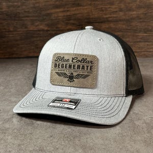May include: A heather gray and black trucker hat featuring a rectangular tan patch on the front. The patch displays the text Blue Collar DEGENERATE, STILL WORKING, STILL DIRTY, STILL BROKE, and an eagle graphic. A black sticker is on the brim.