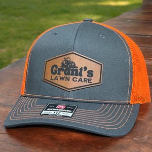 May include: A charcoal gray trucker hat with bright orange mesh back panels and orange stitching. A brown leather-like patch on the front features a lawn mower graphic and the text Grant's LAWN CARE. A black Richardson 112 sticker is on the brim.