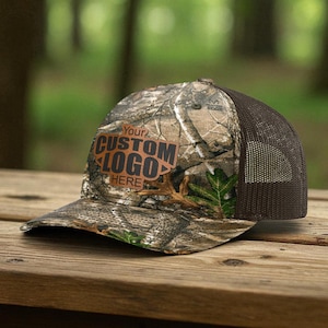 May include: A camouflage print baseball cap with a brown mesh back. The front panel features a brown rectangular patch with the text YOUR CUSTOM LOGO HERE printed in black. The cap has a curved brim and a structured crown with a forest pattern.