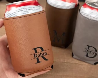 May include: Three personalized can coolers in brown, dark brown, and gray. Each cooler has a name engraved in a bold font. The brown cooler has the name "Robin", the dark brown cooler has the initials "M.A.T", and the gray cooler has the name "Darren".