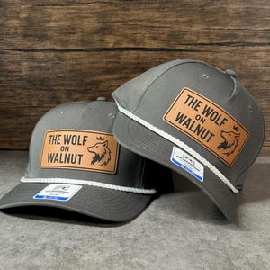 May include: Two grey baseball caps with a white rope detail across the brim. Each cap features a rectangular tan leather patch with the text THE WOLF ON WALNUT and a wolf head graphic. A small white and blue brand sticker is on the brim.