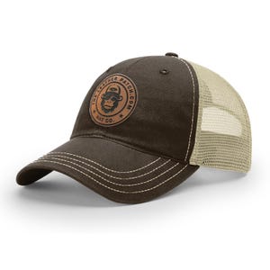 May include: A dark brown baseball cap with a beige mesh back and a curved brim featuring white contrast stitching. A circular tan leather patch is centered on the front, displaying a monkey graphic and the text THE LEATHER PATCH.COM HAT CO.