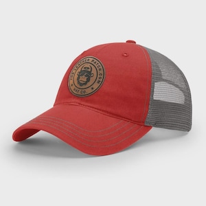May include: A red baseball cap with a grey mesh back and a circular brown leather patch on the front. The patch features a graphic of a gorilla wearing a hat and the text THE LEATHER PATCH.COM HAT CO. The brim has four rows of stitching.