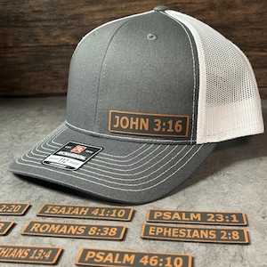 May include: A charcoal gray and white trucker hat featuring a rectangular brown leather patch with the text JOHN 3:16. The hat has a structured front, a mesh back, and a sticker on the brim. Several additional leather patches with Bible verses are displayed.