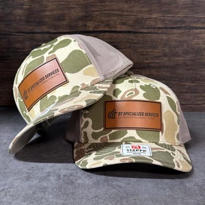 May include: Two camouflage trucker hats with brown leather patches. The patches display the text "DT SPECIALIZED SERVICES" and "ENVIRONMENTAL-DEMOLITION-CIVIL". The hats have a mesh back and a curved brim. The hats are OSFM.