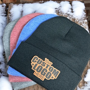 May include: Four knit beanies stacked on top of each other. The top beanie is black and has a brown leather patch with the text "Your Custom Logo Here" in black.