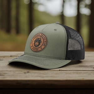 May include: An olive green baseball cap with a black mesh back and a circular brown leather patch on the front. The patch features a graphic of a gorilla wearing a hat and the text THE LEATHER PATCH.COM HAT CO. The cap has black stitching.