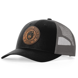 May include: A black baseball cap with a grey mesh back and a circular tan leather patch on the front. The patch features a graphic of a monkey wearing a hat and the text THE LEATHER PATCH.COM HAT CO. The cap has a curved brim and a structured crown.