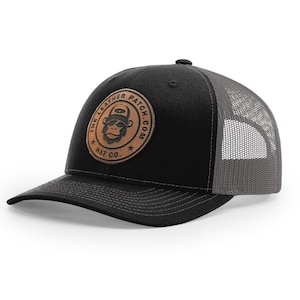May include: A black baseball cap with a grey mesh back and a circular tan leather patch on the front. The patch features a graphic of a gorilla wearing a hat and sunglasses, with the text THE LEATHER PATCH.COM HAT CO. printed around it.