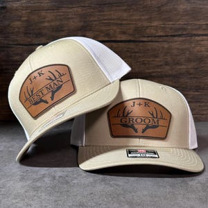 May include: Two beige and white trucker hats featuring brown leather-like patches. One patch displays the text J+K and BEST MAN with an antler graphic, while the other shows J+K and GROOM with an antler graphic. Both hats have a structured front.