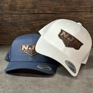May include: Two snapback trucker hats, one in navy blue and one in white, each featuring a brown leather-like patch on the front with the text NJM INDUSTRIES and a small star. Both hats have a mesh back and a circular sticker on the brim.