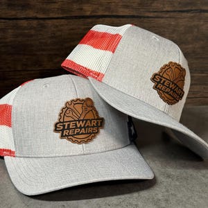 May include: Two heather gray baseball caps with white and red mesh back panels. Each cap features a brown leather-like patch on the front with a gear icon, wrench graphics, and the text "STEWART REPAIRS" embossed in black lettering.