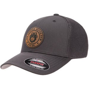 May include: A charcoal gray baseball cap with a mesh back and a circular tan leather patch on the front. The patch features a monkey graphic and the text THE LEATHER PATCH.COM HAT CO. A black sticker on the brim reads THE ORIGINAL FLEXFIT.