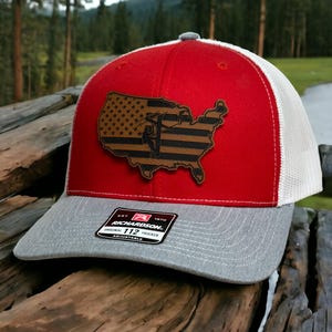 May include: A red and white trucker hat with a grey brim. The front features a brown leather patch shaped like the United States map with a flag pattern and a lineman silhouette. A sticker on the brim reads Richardson Original 112 Trucker Adjustable.