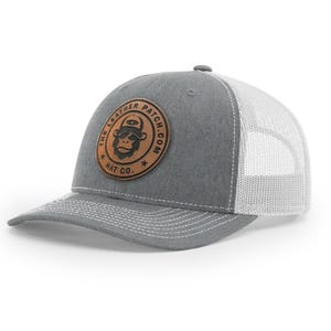 May include: A gray baseball cap with a white mesh back and a brown circular leather patch on the front. The patch features a black graphic of a monkey wearing sunglasses and a hat, with the text THE LEATHER PATCH.COM HAT CO. around it.
