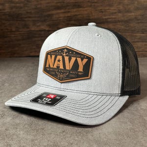 May include: A heather gray trucker hat with a black mesh back and a black brim featuring dark stitching. A hexagonal brown leather patch on the front displays the text NAVY, UNITED STATES NAVY, and an anchor icon with the words FORGED BY THE SEA.