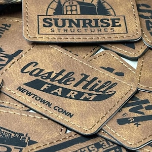 May include: A collection of brown leather patches with various black text designs. One patch reads "Sunrise Structures" with a barn graphic. Another says "Castle Hill Farm, Newtown, Conn". Other patches have sports and business logos.