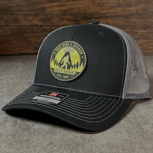 May include: A black trucker hat with a gray mesh back and a curved brim. The hat features a circular patch with the text "PRO HIDE & SEEKER" and an image of a UFO, Bigfoot, and trees. The patch also reads "AREA 51 DIVISION EST. 1967".