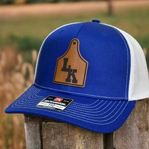 May include: A royal blue and white trucker hat featuring a tan leather-like patch shaped like a livestock ear tag. The patch displays the black letters LK. A black and white sticker on the brim reads Richardson 112 Adjustable.