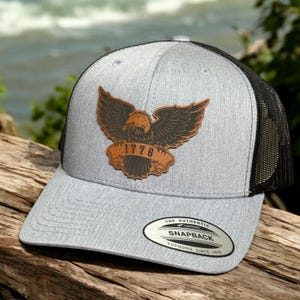 May include: A heather gray baseball cap with a black mesh back and a snapback closure. The front features a brown leather-like patch with an eagle graphic and the text 1776. A circular silver and black sticker is affixed to the flat brim.