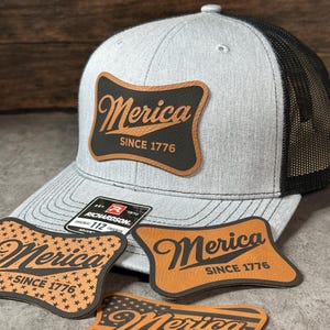 May include: A light gray trucker hat with a black mesh back and a curved brim. A leather-like patch on the front features the text Merica SINCE 1776. Additional patches with the same text and star patterns are displayed in front of the hat.