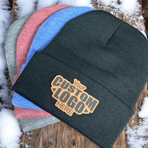 May include: A stack of four knit beanies in black, blue, pink, and gray. The black beanie in the front features a rectangular tan leather patch with the text Your CUSTOM LOGO HERE embossed in black, centered on the folded cuff of the hat.