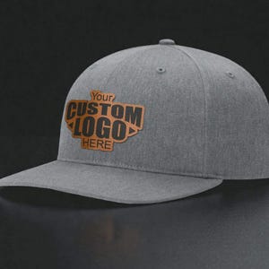 May include: A heather gray baseball cap with a flat brim and a brown leather-like patch centered on the front. The patch features the text "Your CUSTOM LOGO HERE" embossed in a dark, bold font, with a small decorative arrow design on each side.