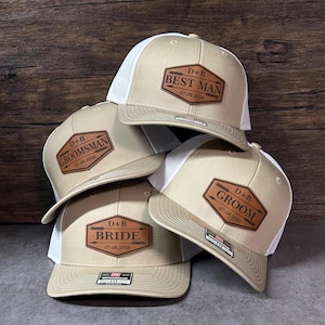 May include: A stack of tan and white trucker hats with leatherette patches. Each hat has text that reads "BEST MAN", "GROOMSMAN", "BRIDE", and "GROOM", along with the initials "D+R" and the date "07.08.2026".