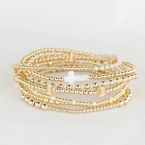 May include: A collection of gold beaded bracelets featuring a white cross charm. The bracelets, composed of small, round gold beads, are layered. The cross charm is crafted from a white, iridescent material.