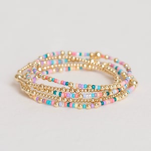 May include: A set of five gold beaded bracelets with colorful beads in shades of pink, purple, teal, and yellow.