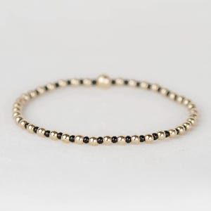 May include: Gold beaded bracelet with alternating gold and black beads. The bracelet is made of small, round beads strung together to form a flexible band. The bracelet is shown on a white surface.