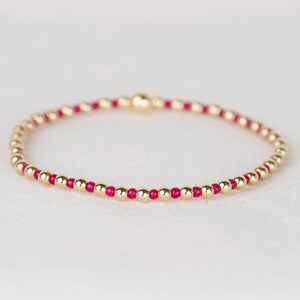 Peut inclure: Un bracelet délicat en perles dorées avec des perles dorées et magenta alternées. Le bracelet est monté sur un bracelet souple, créant un design simple mais élégant. Les perles sont petites et espacées uniformément.