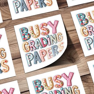 May include: White stickers with the text "BUSY GRADING PAPERS" in colorful, patterned letters. The letters have floral, plaid, and polka dot designs. The stickers are arranged on a wooden surface.