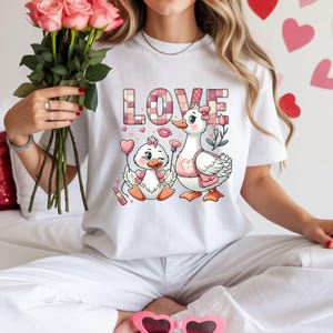 Puede incluir: Camiseta blanca con un diseño de familia de patos de dibujos animados y la palabra "LOVE" en un patrón de cuadros rosas. Los patos tienen accesorios de corazón. La persona sostiene un ramo de rosas rosas.