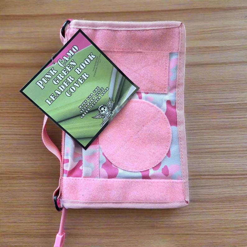 Pu&ograve; includere: Copertina per libro leader rosa mimetico con una toppa circolare rosa e un'etichetta che recita "Pink Camo Green Leader Book Cover". La copertina ha un cinturino rosa e un motivo mimetico rosa e bianco.