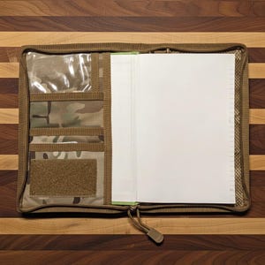 Pu&ograve; includere: Un raccoglitore con cerniera beige e mimetico con pi&ugrave; tasche e una finestra trasparente. Il raccoglitore &egrave; aperto, rivelando una pila di carta bianca a righe. La cerniera &egrave; beige.