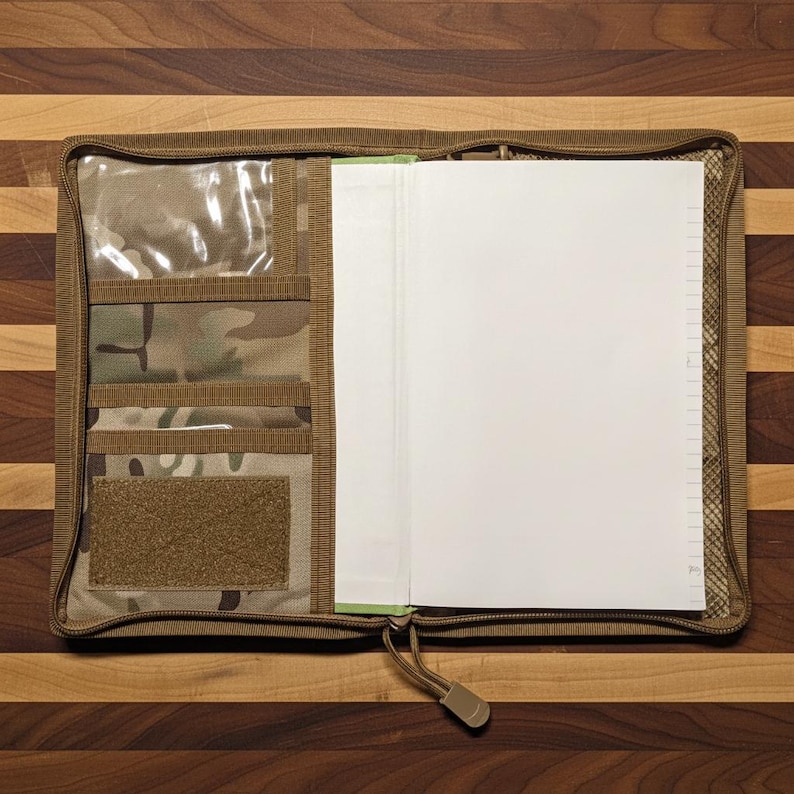 Pu&ograve; includere: Un raccoglitore con cerniera beige e mimetico con pi&ugrave; tasche e una finestra trasparente. Il raccoglitore &egrave; aperto, rivelando una pila di carta bianca a righe. La cerniera &egrave; beige.