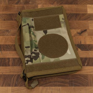 Pu&ograve; includere: Una custodia tattica per notebook con motivo mimetico, caratterizzata da un'ampia toppa circolare e aree rettangolari con chiusura a strappo. La custodia ha una tracolla e una chiusura a cerniera. La tavolozza dei colori include sfumature di verde, marrone e beige.