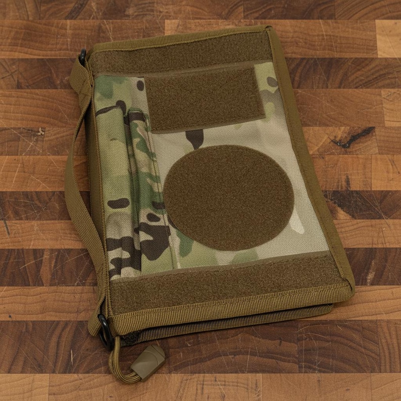 Pu&ograve; includere: Una custodia tattica per notebook con motivo mimetico, caratterizzata da un'ampia toppa circolare e aree rettangolari con chiusura a strappo. La custodia ha una tracolla e una chiusura a cerniera. La tavolozza dei colori include sfumature di verde, marrone e beige.
