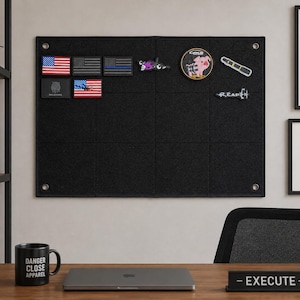May include: A rectangular black felt display board with metal grommets in the corners, featuring various tactical patches including American flags, a pig graphic, and text reading REAPER, mounted on a wall above a desk with a laptop and mug.