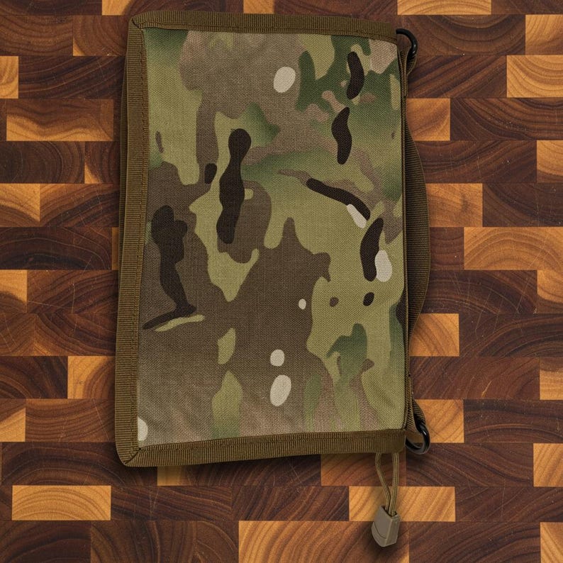 Pu&ograve; includere: Una copertina per notebook rettangolare con motivo mimetico, bordo marrone e cinturino. La copertina presenta un mix di colori verdi, marroni e beige. Un piccolo cordino con un bottone &egrave; attaccato in basso.