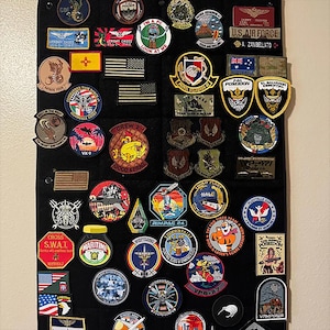 May include: A collection of embroidered patches, including military and aviation-themed designs, displayed on a black fabric. The patches feature various colors, shapes, and text, such as "U.S. AIR FORCE" and "POSEIDON".
