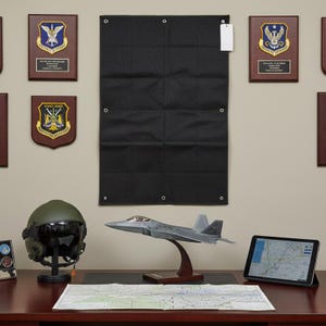 May include: A desk display featuring a gray F-22 Raptor model aircraft on a wooden stand, an olive green flight helmet on a stand, a tablet showing a digital map, a paper map, and a black fabric wall hanging with metal grommets and plaques.