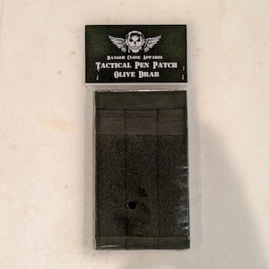 May include: An olive drab tactical pen patch in a clear plastic package. The patch has three pen slots and a hook and loop backing. The top of the package has the words "DANGER CLOSE APPAREL" above a skull and wings graphic, and "TACTICAL PEN PATCH OLIVE DRAB" below.